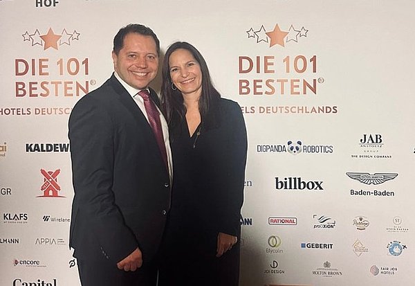 Zwei Personen posieren vor der Sponsorenwand der Preisverleihung „Die 101 besten Hotels Deutschlands“